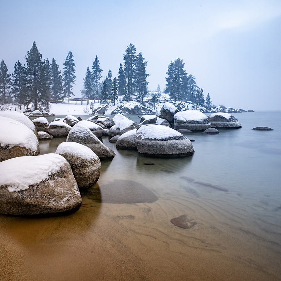Lake shoreline with large boulders and tall trees in winter.
