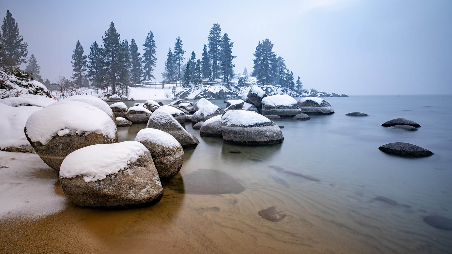 Lakeshore in winter with large snow-covered rocks, tall trees, and boardwalk.