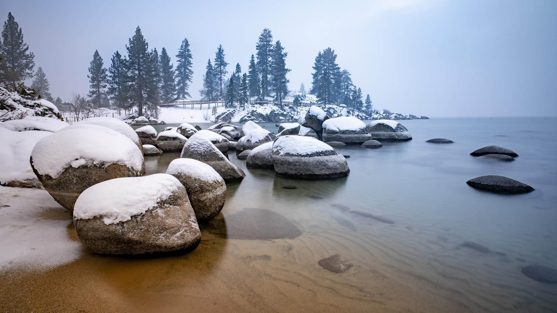 Lakeshore in winter with large snow-covered rocks, tall trees, and boardwalk.