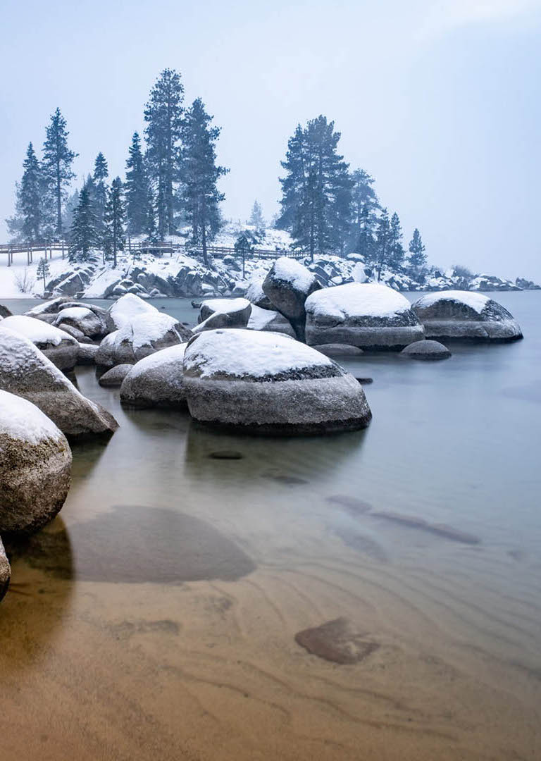 Lakeshore in winter with large snow-covered rocks, tall trees, and boardwalk.